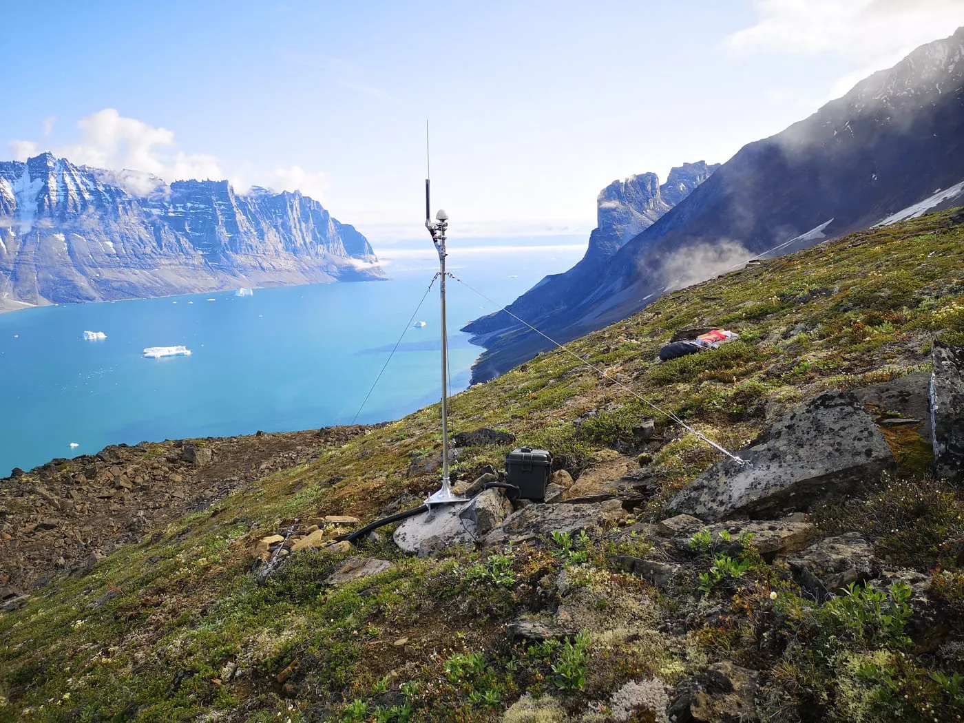 A Seismicstream monitoring station on a grassy slope high above a fjord with drifting icebergs